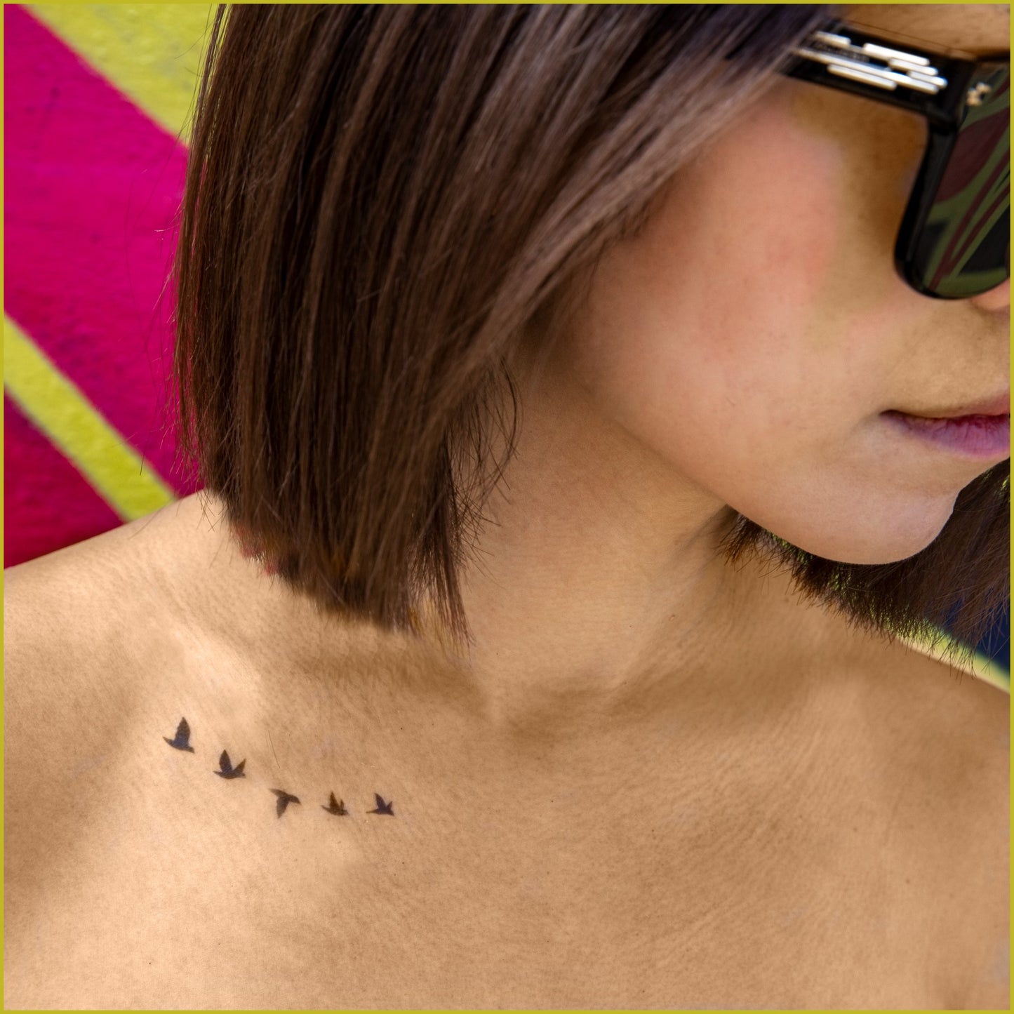 There is a woman with sunglasses in front of a red and yellow graffiti wall. There is a tattoo of a flock of birds below her collar bone.