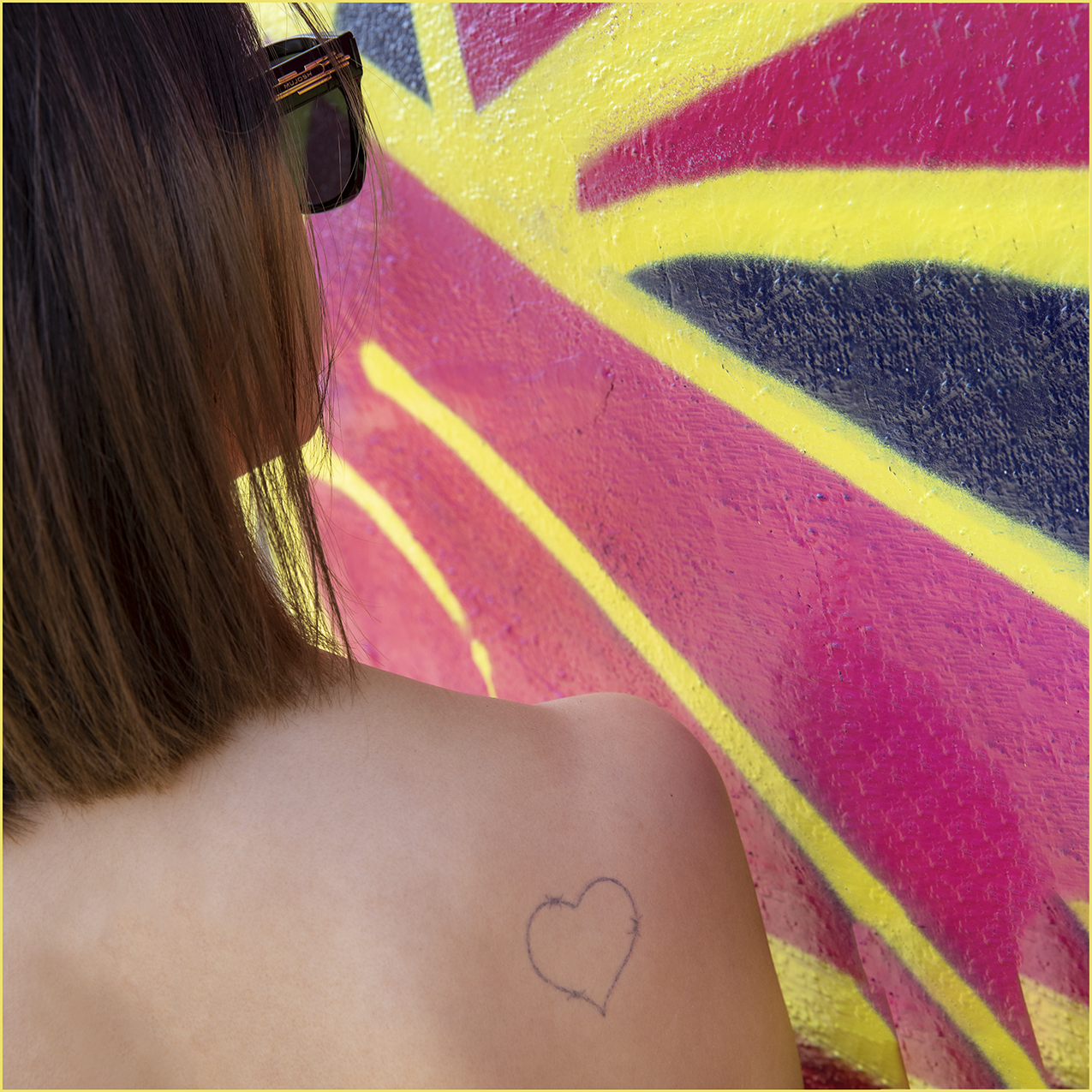 There is a woman in front of a red, yellow and blue graffiti wall, facing the wall. There is a tattoo that represents a barb wired love heart on her right upper back.