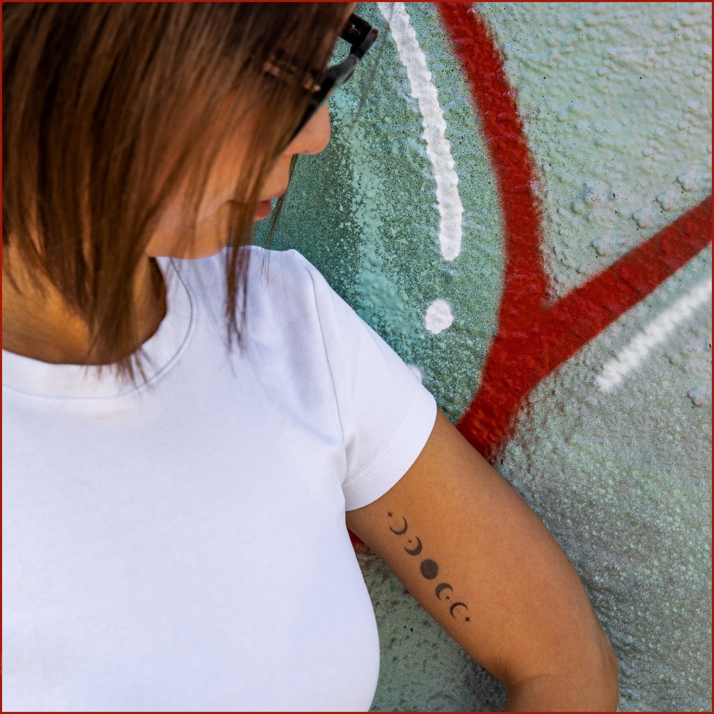 There is a woman in front of a red and green graffiti wall, looking down on her arm. There is a tattoo that represents a line of full moon and half moons on her inner upper arm.