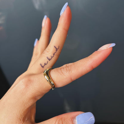 A small tattoo on the side of a middle finger, which reads "be kind". The nails are painted baby blue