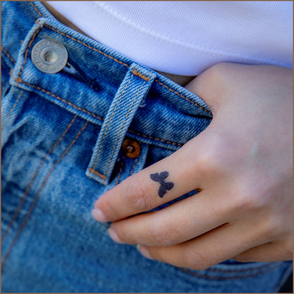 A small tattoo of a butterfly on an index finger of a hand. The hand is resting over denim jeans