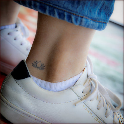 A small tattoo of a lotus on a person's outer wrist. The person is wearing white leather shoes and blue denim jeans