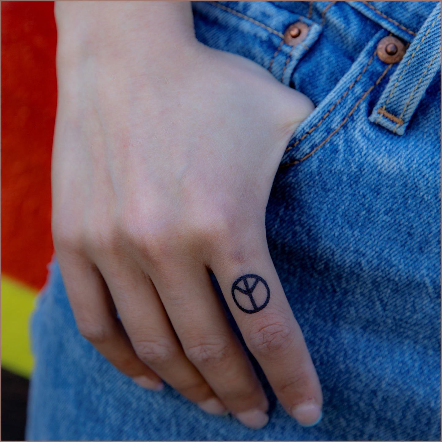 A small tattoo of a peace sign on an index finger of a hand. The hand is resting over a pocket of denim jeans with its thumb in the pocket